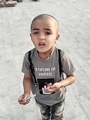 A young boy with a shaved head and some small marks on his face is shown standing outdoors on a light-colored, paved surface, possibly a rooftop or a cleared open area. In the background, a few red bricks are visible. The boy is wearing a grey t-shirt that reads "TO EXPLORE THE UNIVERSE" and has a small black pouch attached to the front. He is also wearing jeans. In his hands, he holds a small, light-colored object with a string attached. He looks directly at the camera, initially with a serious expression, which then transitions into a wide smile as he makes a slight gesture with the object and string. The scene appears to be taking place during the daytime under bright, clear weather conditions in Dera Chahal, Pakistan.