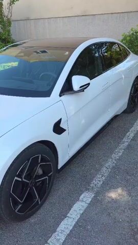 A white BYD sedan is parked on an asphalt surface. The car has a black roof and black accents on the wheels and front fender. The camera pans across the front of the car, showing the headlights and the BYD logo on the hood. The surrounding environment includes a building and some greenery.