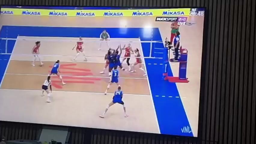 A television screen, viewed from Xàtiva, Spain, displays a live indoor women's volleyball match in progress. The wide shot captures most of the court, showing players from two teams. One team wears blue uniforms, while the opposing team is in red and white. There are at least ten players visible on the court, actively engaged in the game. The ball appears to be in play, with players positioned to receive or attack. A referee stands on a high chair next to the net, overseeing the match.

The court itself is marked with blue lines and a prominent brown area, featuring large red arrow-like designs. Banners around the court's perimeter and on the net feature the logo "BANCO DO BRASIL." Text on the court's sideline also reads "BRASILIA BRAZIL," indicating the event's location. A digital overlay on the TV screen shows "MAXSPORT 2HD LIVE" along with a timestamp or score of "23:48". The overall atmosphere suggests a competitive and focused sporting event. A "JNL" watermark is visible in the bottom right corner of the TV screen.