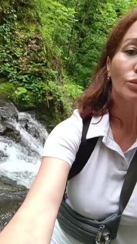 A woman with reddish-brown hair and fair skin is taking a selfie video while standing near a waterfall. She is wearing a white polo shirt, a black backpack, and a gray fanny pack. She is also wearing light-colored pants and black sandals. The waterfall is surrounded by lush green foliage. The woman is smiling at the camera.