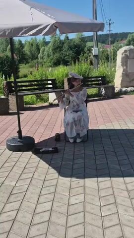 A young woman in a long, white dress with a floral pattern and a hat is playing a flute while seated outdoors. She is positioned under a large, light-colored umbrella, which casts a shadow on the brick-paved ground. To her left, a black speaker sits on the ground, and a small black object is positioned near her feet. In the background, there is a wooden bench, greenery, trees, a stone monument, and buildings. The sky is blue and sunny. Several people can be seen walking in the distance.