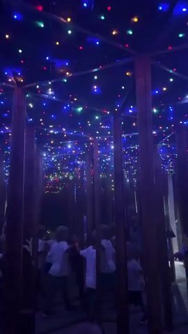 The video shows a group of people, including several children and adults, walking through a mirror maze. The maze is decorated with numerous colorful LED lights on the ceiling, creating a dazzling effect. The walls of the maze appear to be made of mirrors, reflecting the lights and the people within. Wooden posts are also visible, dividing the paths of the maze. The lighting is dim, with the colorful lights being the primary illumination. The people are casually dressed and seem to be enjoying themselves as they navigate the labyrinth. Some are looking around, while others are interacting with each other. The overall atmosphere is playful and festive.