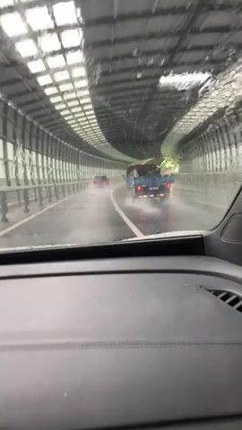 The video is taken from the interior of a car, looking through the windshield at a tunnel. It is raining heavily, and the windshield is wet and streaked. Several vehicles are visible in the tunnel, including a red car ahead, a blue truck to the right, and other vehicles further in the distance. The tunnel walls are lined with sound-absorbing panels, and there are green signs indicating exits. The road is wet and reflects the lights of the vehicles and the tunnel. The video captures the perspective of a driver navigating through the tunnel during a downpour.