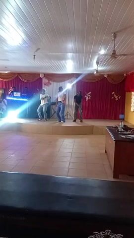 Three people dance on a small stage in front of a red and gold curtain. One person wears a white shirt and jeans with a white hat, one person wears a white shirt with wide-leg jeans and a pink waist bag, and one person wears a black shirt and patterned cargo pants. There is a DJ behind them with blue lights. There is a table with drinks on it to the right of the stage. The ceiling has white panels and two ceiling fans. The floor is made of light tiles. The camera slowly pans from the floor to focus on the stage.
