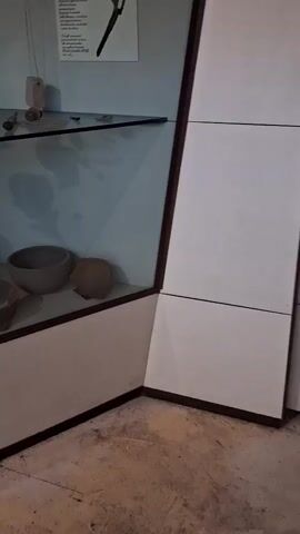 A museum display case contains ancient pottery. The pottery is on shelves behind glass. The display case is lit from above. The pottery is various sizes and shapes. Some pieces are broken. There is a text label next to one of the pottery pieces. The text is in Italian. The pottery is made of terracotta. The display case is made of wood and glass. The floor is tiled.