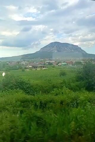 The video shows a view from a train window of a rural landscape. In the foreground, there is lush green grass and some bushes. In the middle ground, there are several houses with red roofs, some fences, and dirt roads. In the background, there are rolling hills and a large, conical mountain under a cloudy sky. The train appears to be moving, as the landscape is subtly shifting.