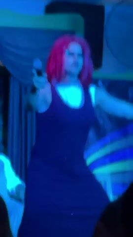 The video shows a person on a stage, likely performing. The person is wearing a wig with bright red hair, sunglasses, and a dark dress over a white shirt. They are holding a microphone and appear to be singing or speaking. The stage is lit with colorful lights, including blue, pink, and red, creating a dynamic and theatrical atmosphere. In the background, there is a fan and some draped fabric. The audience is partially visible in the foreground, with their faces obscured. The overall scene suggests a live performance, possibly a drag show or a concert.