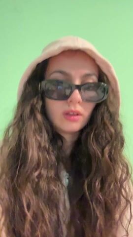 A woman with long, curly brown hair, wearing a light pink hooded sweatshirt and dark sunglasses, is facing the camera. She is sitting against a brown surface that appears to be a sofa, in front of a pale green wall. She covers her mouth with her hand, then looks at her fingers, displaying a manicure with alternating yellow and red nails. She then touches her chest with the same hand. The woman appears to be unwell.