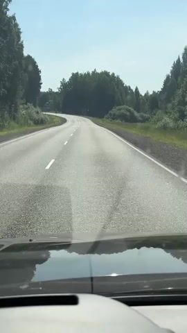 The video is a first-person perspective from a car driving on a paved road. The road is in good condition with white lane markings. The scenery on either side of the road is green with trees and vegetation. The sky is clear and blue, indicating a sunny day. The road curves gently to the left as the car continues to move forward. There are no other vehicles or people visible in the frame.