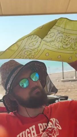 A man wearing a straw hat, sunglasses, and a red t-shirt is sitting under a light-colored awning at a beach. He is holding a yellow bandana above his head with his right hand, tilting his head back to look at it. The bandana has a patterned design. In the background, there is a beach with sand, some lounge chairs, and the ocean. The sky is clear and blue.