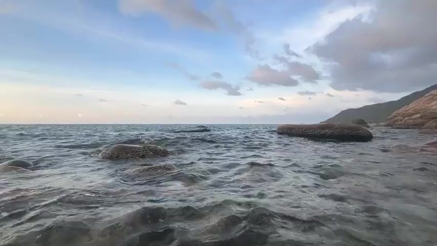 The video shows a wide shot of a rocky beach. The camera is positioned low to the ground, capturing the water washing over the rocks near the shore. Several large rocks are partially submerged in the water. The water is clear and relatively calm, with small waves rippling across the surface. In the background, the horizon meets a partly cloudy sky with shades of blue and grey. A portion of a hill or landmass is visible to the right of the frame.