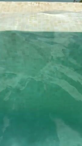 The video opens with a close-up shot of the edge of a swimming pool. The water is clear and greenish, with tiles visible beneath the surface. The edge of the pool is lined with small, square, light-colored tiles. The camera then pans up and away from the pool, revealing a vast, turquoise lake or reservoir in the background. The lake is surrounded by rolling, arid hills and dotted with trees. The sky is partly cloudy, with dramatic, white clouds against a blue backdrop. The overall impression is one of a serene, picturesque landscape. The video appears to be filmed during the daytime. No people are visible in the video.