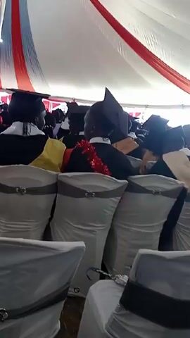 The video shows a large group of people seated in white chairs under a large white tent with red and blue stripes. Most of the individuals are wearing graduation gowns and mortarboards, indicating a graduation ceremony. The view is from behind the attendees, focusing on the back of their heads and shoulders. The chairs are arranged in rows, and the overall atmosphere suggests a formal event.