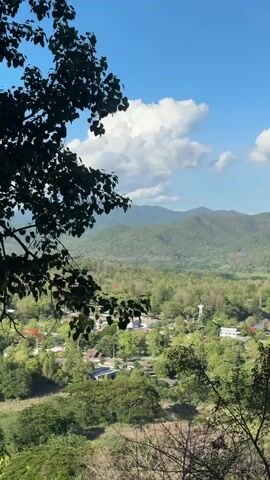 The video shows a scenic view of a small town nestled in a valley, surrounded by lush greenery and mountains in the background. The sky is blue with fluffy white clouds. The camera is static, framing the landscape from a slightly elevated position. The foreground features tree branches and foliage, partially obscuring the view. In the middle ground, there are houses and buildings scattered among the trees, and a lake or river can be seen winding through the valley. The mountains in the background are covered in dense forest, creating a picturesque backdrop. The overall impression is one of peace and natural beauty.