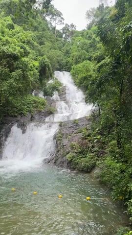 A vertical shot captures a multi-tiered waterfall cascading down a rocky, tree-covered mountainside. The water flows over gray rocks and lush green foliage, creating a frothy white torrent that collects in a clear, calm pool at the bottom. Yellow buoys float in the pool, suggesting it is a designated swimming area. The surrounding environment is dense with green trees and vegetation, indicating a natural, possibly tropical, setting. The sky is overcast.