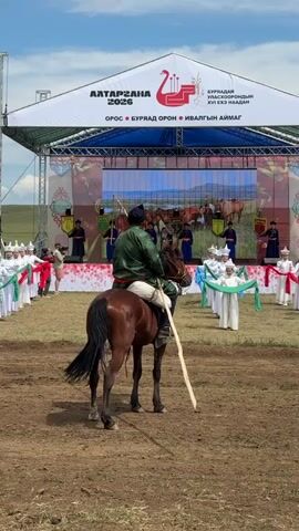 The video shows a horse and rider positioned in the foreground, facing away from the camera. The rider is wearing a traditional green garment and a dark hat, holding a long stick. The horse is brown with a dark mane and tail. Behind the horse and rider, a stage is set up with a large white banner overhead. The banner displays text in Mongolian and the year 2026, along with a graphic of a bird. The stage itself features a large screen displaying an image of horses and a landscape. Several people are standing on the stage, some wearing traditional white and green attire, holding long green ribbons. On either side of the stage, rows of people in similar traditional outfits are standing. The ground in the foreground is dirt and grass. The sky is blue with some clouds. The overall scene appears to be an outdoor cultural event.