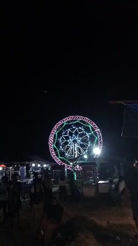 A Ferris wheel is illuminated at night with multicolored lights. People are gathered around stalls selling various items. The scene appears to be a fair or festival.