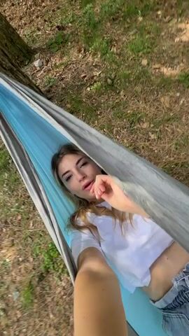 A young woman with long brown hair, wearing a white crop top and blue denim shorts, lies in a blue and gray hammock. She holds a camera in her right hand, capturing a selfie shot from a high angle. She briefly glances down at the camera, then looks up and smiles. She then swings the camera down towards the ground and then back up to her feet, which are also in the hammock. The hammock is tied to a large tree, and the background shows a forest with tall trees and green grass.