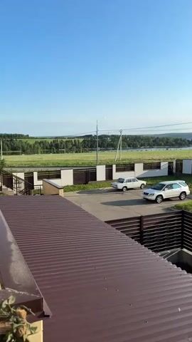 The video shows an outdoor scene with a clear blue sky and distant trees and water. In the foreground, there is a brown metal roof. Behind that, a brown slatted fence encloses a small, overgrown area. Further back, a paved area has two cars parked on it, one white SUV and one white sedan. Beyond the paved area, another fence with white panels and brown slats runs horizontally across the frame. Beyond this fence, there is a grassy field with some trees and a body of water in the distance. There are utility poles with wires visible in the background. The lighting suggests it is daytime, likely in the morning or afternoon. There are no people visible in the video.