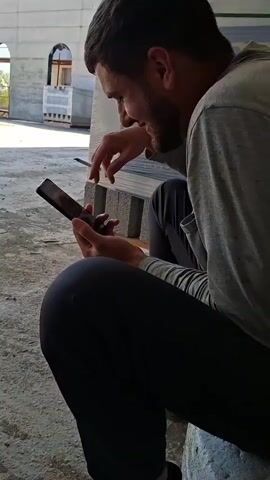A man with a beard and short brown hair is sitting on a rough surface, possibly a concrete ledge. He is wearing a gray long-sleeved shirt and dark pants. He is looking down at a black smartphone he holds in his left hand, while his right hand appears to be operating the screen. In the background, there is a partially constructed building with concrete walls and arched openings. There are also some stacks of what appear to be construction materials, including gray blocks and pallets. The lighting suggests it is daytime.