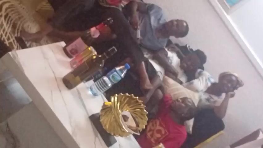 The video shows a group of people sitting close together in a room.  There are six individuals, including one child.  The others are adults, with at least two women and one man visible. They appear to be looking at the camera and smiling.  A white, marble-like table is positioned in front of them. Several bottles, including alcohol bottles and a water bottle, are on the table. There is also a gold decorative object. The room appears to be a living room, with a rug and a picture frame visible in the background. The camera is slightly angled, providing a top-down view of the scene.