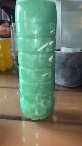 A full bottle filled with green liquid sits on a wooden table. There is some white residue on the bottle, likely from a label. In the background is a bottle of Tango soda and a concrete pot. The lighting is bright.
