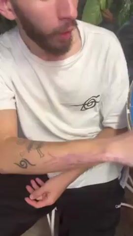 A man in a white t-shirt with a swirl-shaped logo and black sweatpants is sitting on a white plastic chair, extending his left arm forward to show a tattoo on his forearm. The tattoo is purple and appears to be multiple scratch marks. There are two additional tattoos visible on his upper arm: one is a cartoon character with a tribal symbol below it, and the other tattoo is a tribal symbol alone. He has a beard and is looking off camera as he shows the tattoo. A wet floor sign is visible in the background.