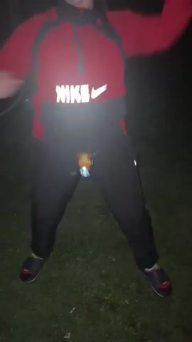 A person in a red Nike hoodie and black pants with a headlamp attached to their waist is jumping and dancing in a grassy area at night. The headlamp flashes with each movement. The person's face is not visible. The background is dark and out of focus.