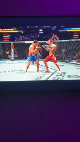 A screen displays a UFC video game in progress. The characters in the game are inside the ring. One character wears blue trunks and the other red. The character in blue throws a punch, and the character in red falls to the ground. Then the character in blue goes back in to fight. He goes low and the character in red delivers a kick. The surrounding environment shows a wall behind the TV.