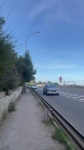 The video shows a roadside scene with a sidewalk on the left and a multi-lane road on the right. Cars are driving on the road. A pedestrian sidewalk is visible on the left side, with a low wall and vegetation lining it. A street lamp extends over the sidewalk. On the right side, a metal guardrail separates the sidewalk from the road, which has multiple lanes. Several cars are driving on the road, including a silver SUV in the foreground, a white hatchback, a dark sedan, and another dark sedan. A speed limit sign indicating 50 is visible on the right side of the road. The sky is blue with some clouds, suggesting daytime. The environment appears to be an urban or suburban area.