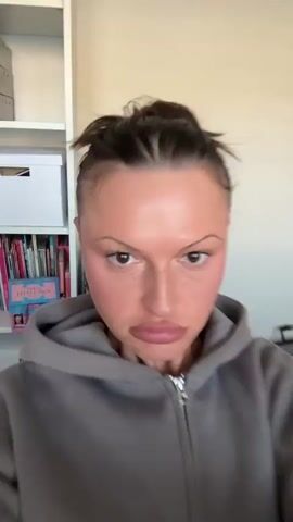 The video features a close-up of a woman's face in an indoor setting. She has fair skin with numerous small freckles visible across her nose and cheeks, dark brown eyes, and styled eyebrows with distinct winged eyeliner. Her lips are full and accentuated with a matte pinkish-brown lipstick. Her hair is pulled back, and she is wearing a grey hooded sweatshirt.

The video comprises three short segments. In the first clip (0-1 seconds), the woman's face is shown frontally in a very tight shot. She exhibits a serious or pensive expression, with a slight furrow in her brow and her mouth slightly open as if about to speak.

The second clip (1-2 seconds) transitions to a profile view, showing her right cheek, ear, and part of her neck. She moves her mouth, suggesting she is speaking or reacting. In the blurred background, a white bookshelf filled with books is partially visible, indicating a residential or private study environment.

The final clip (2-3 seconds) returns to a frontal close-up of her face. She looks directly into the camera with wide, expressive eyes and a subtle smile, conveying a playful or perhaps mischievous emotion.

The lighting throughout the video is bright and even, consistent with an indoor setting during daylight hours. No specific time of day or weather conditions are discernible, other than the indoor context. No visible text is present in the video. The location context provided indicates this scene is taking place in Tunbridge Wells, United Kingdom.