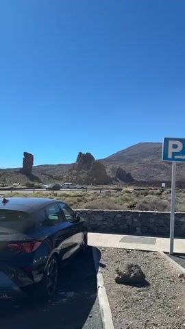 A vertical video pans across a parking lot under a clear blue sky. The parking lot is bordered by a low stone wall on the left and asphalt on the right. A blue and white parking sign with a car icon is visible. Several cars are parked in designated spots. In the background, a volcanic landscape with a large mountain dominates the view, with a small building resembling a church or visitor center situated to the right. Further down the road, more buildings and a bus are visible, with a group of people gathered. The video pans from left to right, starting with a view of a dark-colored car and then revealing the wider landscape and parking area.
