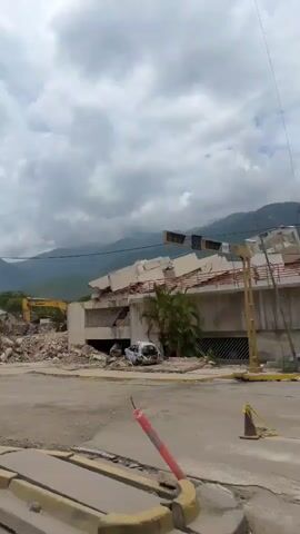 A low-angle, wide shot shows a scene of destruction from an earthquake. In the foreground, a street is littered with debris. A yellow excavator is visible in the middle ground, partially obscured by rubble. To the right of the excavator, a partially collapsed building stands, with a white car visible amongst the debris. The background features mountains under a cloudy sky. The video appears to be taken from a moving vehicle, as indicated by the slight camera shake.