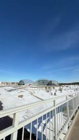 The video opens with a static, wide shot of a snow-covered landscape under a bright blue sky, featuring a distinctively shaped building in the distance. The camera gradually transitions, moving along a pedestrian bridge with white metal railings. The view shifts to follow a woman pushing a stroller with a young child inside across the bridge, a man walking alongside. The scene is brightly lit, with the sun prominently visible in the sky. The landscape below the bridge is covered in snow, and the day appears crisp and clear. The woman is wearing sunglasses and a brown jacket, and the man is dressed in a black jacket.