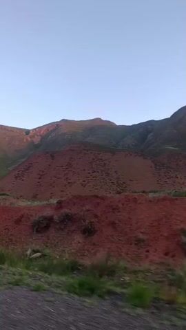 The video shows a landscape with mountains in the background and a red, barren hill in the mid-ground. In the foreground, there is a dirt road and some green vegetation. Traffic signs are visible on the left side of the road. The sky is clear and blue. The scene appears to be a rural or remote area. The sun is low in the sky, suggesting it is either sunrise or sunset, casting a warm glow on the mountains.
