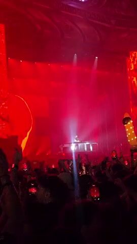 The video shows a crowded concert with a DJ performing on a stage. The stage is lit with red lights and smoke. The crowd is holding up their phones to record the performance. In the background, large screens display images of people. The overall atmosphere is energetic and exciting.