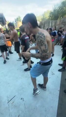 A crowd of people are gathered outdoors. Many are wearing casual clothing, such as t-shirts and shorts. Some individuals are shirtless. The environment appears to be a paved area with a rocky, textured wall in the background. The lighting suggests it is daytime. Some people are dancing or moving to music. One person in the foreground is shirtless and wearing light blue shorts and black and green sneakers. They have tattoos on their arm and are wearing a black baseball cap. Another person in the background is also shirtless and wearing light blue shorts. The image is from a low angle, looking up slightly at the people.