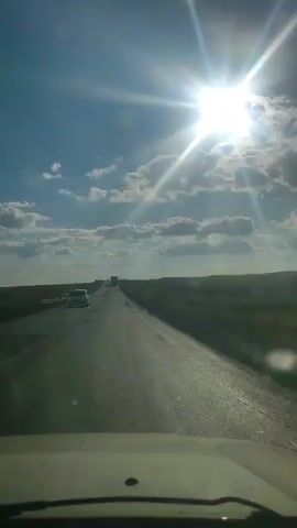 The video shows a point-of-view shot from a vehicle driving on a road under a bright, sunny sky with scattered clouds. The road is asphalt and appears to have some potholes. To the left, there are guardrails and flat, grassy terrain. To the right, the terrain is also flat and grassy. There are a few other vehicles visible in the distance on the road, including what appears to be a truck. The sun is shining brightly, creating lens flares in the video.