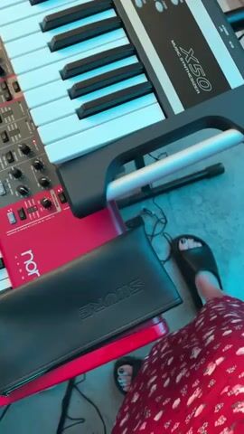 A close-up shot captures two stacked electronic keyboards, a black Korg X50 Music Synthesizer on top of a red Nord Lead 2x, positioned in a dark environment with blue and green lighting. The keys of both keyboards are predominantly white with black accents. An iPad, displaying a music-related interface, rests on a stand between the keyboards. A black leather case with "SHURE" written on it is visible on the Nord keyboard. A person's feet, wearing black slide sandals and a red patterned dress, are at the bottom right of the frame, tapping rhythmically.