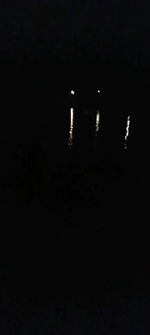 This is a dark video that appears to be filmed at night. The dominant visual element is the reflection of lights on a body of water. These reflections appear as vertical streaks of light, suggesting the presence of lights on the shore or on a boat. The overall scene is very dark, with minimal detail visible beyond the light reflections.
