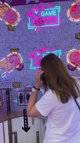The video shows a bright and colorful arcade with neon signs and stuffed animals. Two people, a woman in a white top and shorts, and a man in a white shirt, are walking through the arcade. The woman is in the foreground, and the man is slightly behind and to the right. They are walking past a row of claw machines filled with stuffed animals. The walls are decorated with cartoon characters and neon signs that say "GAME ZONE." In the background, there are more arcade games and a seating area with tables and chairs. The overall atmosphere appears to be fun and lively.