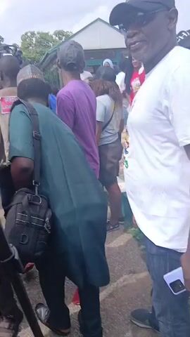 The video shows a crowd of people gathered outdoors. There is a professional video camera on a tripod in the foreground, operated by someone whose arm and shoulder are visible. Several individuals are visible in the background, some wearing t-shirts with political imagery and slogans, suggesting a possible event or rally. The environment appears to be paved, with trees and buildings in the distance under an overcast sky. People are looking in various directions, and some appear to be engaged in conversation or observing something. One person in a purple shirt and black pants with red shoes stands out in the middle ground. Another individual wearing a blue t-shirt and a woman in a blue shirt with braided hair are also noticeable. A man in a white t-shirt with colorful graphics is on the far right. The overall impression is of a public gathering possibly related to a political event or media coverage.