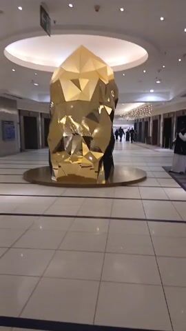 The video shows an interior shot of a large, well-lit space with a high ceiling and a large, geometric gold sculpture in the center. The sculpture appears to be made of flat, angular panels. The floor is tiled in light colors with darker accent lines. In the background, people are walking, including a few individuals dressed in dark clothing. The general atmosphere is that of a public space, possibly a shopping mall or hotel lobby. There are doorways and signs indicating directions. The camera is relatively still.