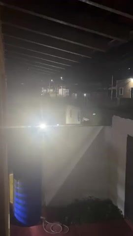 At night, the camera peers out from under a wooden roof structure. Below, a blue water tank with the words "Resinca Tanker 770" is visible. Behind the tank, a white wall separates the area from a dimly lit street. A small patch of green plants grows at the wall's base. In the street, lights from streetlamps and a passing car shine brightly, creating a lens flare effect in the camera. Buildings line the street, and there is a slight haze in the air. The scene is dark and still.