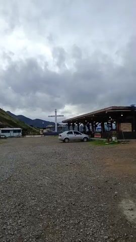 The video shows a landscape with a restaurant, a large cross, and a silver sedan under a cloudy sky. The ground is covered with gravel. Beyond the restaurant is a construction site and a mountainous landscape covered in fog. The sky is overcast, but with glimpses of blue sky appearing behind the clouds. The video pans from the cross and car to the building and then the mountain view.