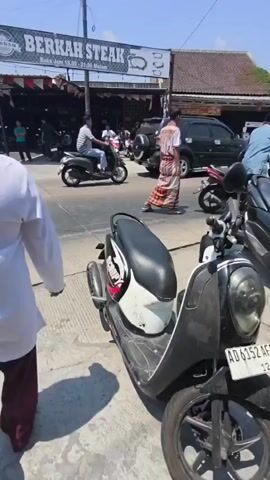 The video shows a street scene on a sunny day. In the foreground, a black and white scooter is parked on the right side of the road. Several other scooters are visible in the background, some with riders. People are walking and riding motorcycles along the street, which appears to be lined with shops or stalls. A sign that reads "BERKAH STEAK" is visible on one of the buildings. One person in the foreground is wearing a white shirt and a checkered sarong. Another person further down the road is also wearing a checkered sarong and a white shirt, and they appear to be walking across the street. There are also a few cars parked on the side of the road. The overall impression is of a busy street in a warm climate, likely in Southeast Asia given the style of clothing and the presence of scooters.