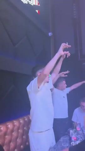 Two young men are dancing with their arms raised in a dimly lit room with blue and purple lighting. Both are wearing white t-shirts. The man in the foreground is wearing glasses and holding a cigarette in his right hand. The man in the background is also holding a cigarette. There is a plush brown sofa to the left of the frame and a table with drinks in front of the men.