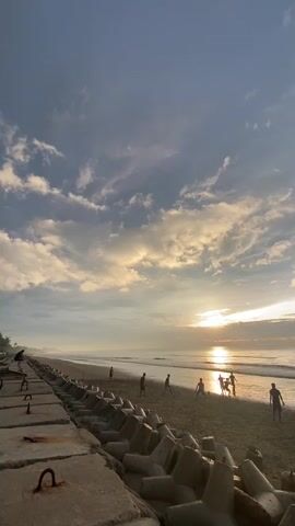 The video shows a beach scene at sunrise. In the foreground, a concrete structure with C-shaped metal pieces is visible, likely a seawall or breakwater. Beyond this, a sandy beach stretches out, with several people playing on it. The sun is rising over the ocean, casting a warm glow on the water and sky, which are dotted with clouds. The ocean appears calm with gentle waves. The overall atmosphere is serene and idyllic.