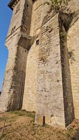 The video begins with a close-up, low-angle shot of a stone tower, possibly part of a castle or old building, against a clear blue sky. Sunlight illuminates the weathered stone and the architectural details, including small windows and crenellations at the top. A sparse tree is growing from a crevice in the tower's wall. As the video progresses, the camera pans slightly downwards and to the right, revealing more of the tower's base and the surrounding grounds. A patch of dry grass and some low-lying vegetation are visible at the bottom of the frame. Further to the right, a dirt path leads away from the tower, and in the distance, a white van and other vehicles are parked. Two people, a man and a woman, are walking away from the camera on this path, both carrying what appear to be rackets. The woman is wearing shorts and a tank top, while the man is wearing shorts and a t-shirt. The background shows a rural landscape with fields and trees under a bright, sunny sky. The scene appears to be outdoors on a warm, clear day.