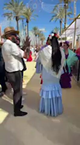 The video captures an outdoor scene at a lively festival in Jerez de la Frontera, Spain, likely during daylight hours given the bright, clear blue sky. Palm trees and white tent structures (possibly casetas) line the area.

The primary subjects are a man and two women, all dressed in traditional Spanish attire. The man, wearing a white shirt, dark trousers, a vest with a strap, and a fedora, claps his hands to a rhythm. He adjusts his hat and looks towards the camera with a subtle smile, indicating a relaxed and enjoyable atmosphere.

Two women are prominently featured. One woman, with long dark hair adorned with flowers, wears a light blue traditional dress with ruffled layers and a white fringed shawl. Her back is mostly to the camera, but she turns and interacts with another woman. This second woman wears a bright red ruffled dress with a white top. They appear to be greeting each other or adjusting their attire, reflecting a communal and celebratory mood.

The overall activity suggests a traditional Spanish festival or fair, characterized by the distinctive clothing and lively atmosphere. People in the background are also visible, further contributing to the festive ambiance. No visible text is clearly discernible in the video.